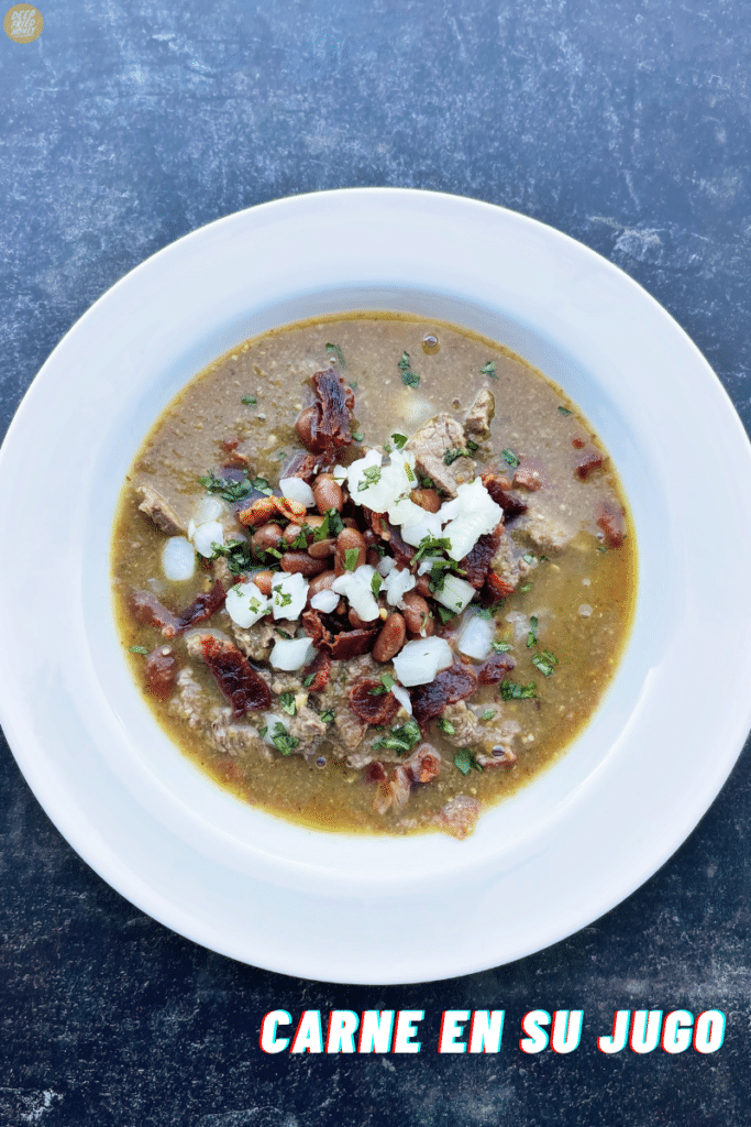 white bowl of Carne en Su Jugo, a mexican stew, topped with bacon, minced cilantro, chopped white onions, and diced crispy bacon.