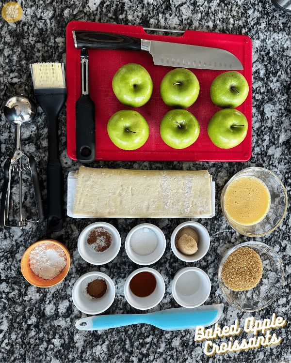 mise en place for recipe: green apples, a peeler and chef's knife on a red cutting board, beside small bowls of sugars and spices, folded puff pastry, egg wash, a cookie scoop, spatula, and basting brush.